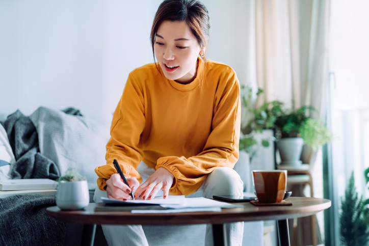 Young Asian woman holding a pen and signing paperwork in the living room at home