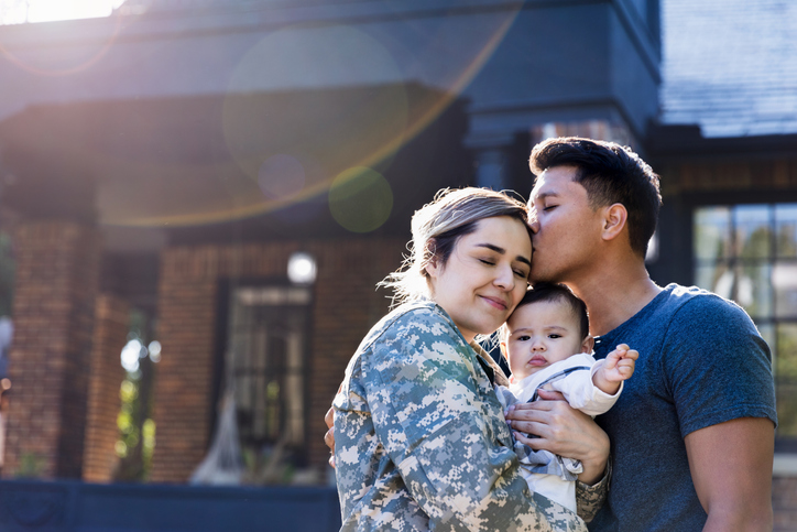 Man kisses his soldier wife holding a baby in front of a home.